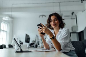 Woman in a white shirt smiling while using a perimenopause symptom tracking app on her phone at a desk