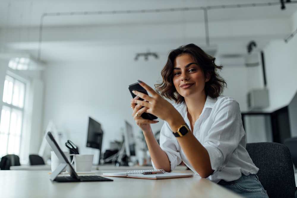 Woman in a white shirt smiling while using a perimenopause symptom tracking app on her phone at a desk
